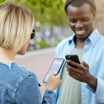 Duas pessoas, um home e mulher utilizando celulares com o olhar para o dispositivo sorrindo.